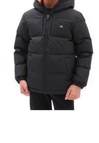 Persona con una chaqueta acolchada negra de Vans, con capucha y cremallera delante, de pie contra un fondo blanco.