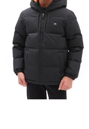 Persona con una chaqueta acolchada negra de Vans, con capucha y cremallera delante, de pie contra un fondo blanco.