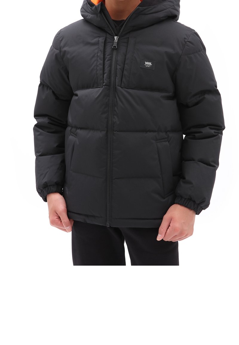 Persona con una chaqueta acolchada negra de Vans, con capucha y cremallera delante, de pie contra un fondo blanco.