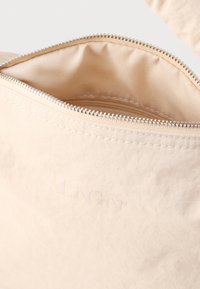 Μπεζ θήκη με φερμουάρ, κατασκευασμένη από μαλακό, υφή υλικό. Διαθέτει ομαλή εσωτερική επένδυση και ανάγλυφο λογότυπο στην εξωτερική πλευρά.