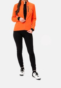 Klar orange långärmad jacka med svarta detaljer och ficka med dragkedja, tillsammans med svarta tajta byxor och vita cykelskor. Slät textur.