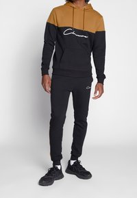 Felpa con cappuccio nera e marrone con logo bianco, abbinata a pantaloni jogger neri con dettagli marroni e un design aderente.