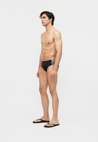 Man zonder shirt, gekleed in zwarte zwemslips met rode en dierenprintpanelen en zwarte teenslippers, staand in profiel tegen een effen witte achtergrond.