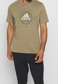 Oliven grøn bomulds t-shirt med et cirkulært grafik med Adidas-logoet i hvid, korte ærmer og rund halsudskæring.
