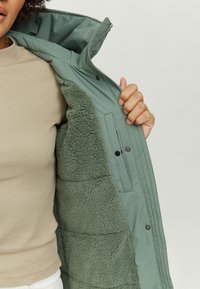 Green padded jacket featuring a soft, fluffy interior lining, zip closure, and snap buttons. The outer material has a smooth texture.