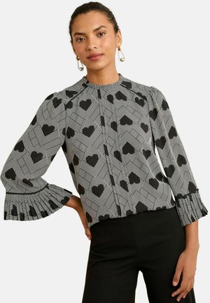Black and white long-sleeve blouse featuring a geometric heart pattern, ruffled cuffs, and a high neckline for a structured design.