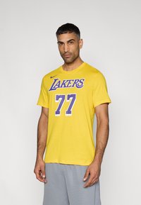 Κίτρινη βαμβακερή μπλούζα με την επιγραφή «LAKERS» σε μωβ γράμματα και τον αριθμό «77» σε λευκό. Με στρογγυλή λαιμόκοψη και κοντά μανίκια, συνδυασμένη με γκρι σορτς.