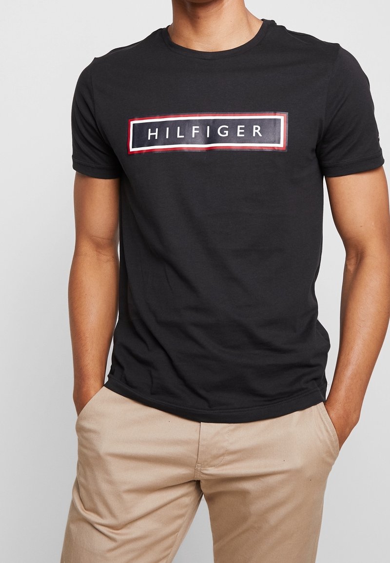 Černé bavlněné tričko s kulatým výstřihem a krátkými rukávy, na kterém je obdélníková grafika s nápisem "HILFIGER" v bílé barvě, obtažená červenou a modrou.