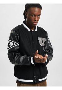 Veste varsity noire avec manches en cuir, col et poignets rayés blancs, ornée de détails décoratifs et d'une fermeture à boutons à l'avant.