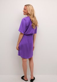 Violeta īsu piedurkņu kleita ar elastīgu jostasvietu, izliekta apakšmala un brīva piegriezuma. Modelis, uzvilcis melnas plakanas sandales, pozē no aizmugures.