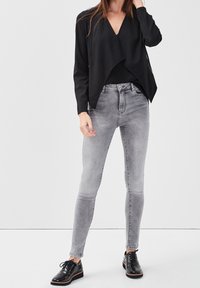 Veste en maille noire avec devant drapé, associée à un jean skinny gris clair et des chaussures en cuir verni noir, présentant une texture lisse.