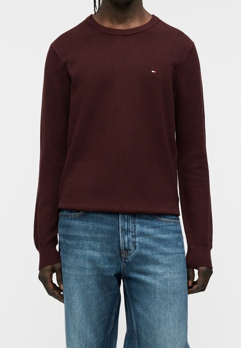Personne portant un pull bordeaux à manches longues avec un petit logo sur la poitrine et un jean bleu, debout devant un fond clair et uni.