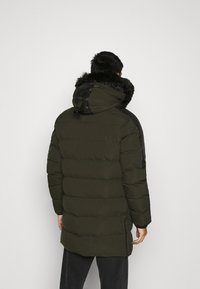 Parka acolchada en verde oscuro con capucha forrada de piel sintética, detalles negros en los hombros y aberturas laterales para facilitar el movimiento. Textura suave.
