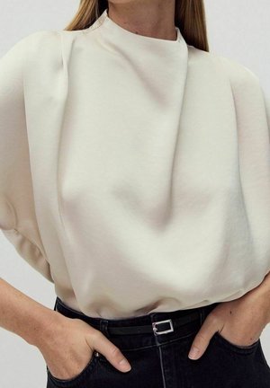 Femme portant un chemisier blanc structuré rentré dans un jean noir taille haute avec une ceinture noire, les mains dans les poches.