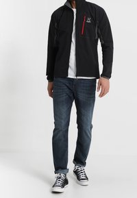 Svart softshell-jacka med grå detaljer och röd dragkedjeficka, bärs över en vit skjorta, i kombination med blå jeans och svarta sneakers.