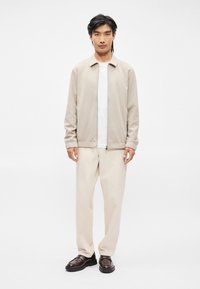 Jasnobeżowa kurtka zapinana na zamek z kołnierzykiem, zestawiona z białą koszulką i kremowymi spodniami. Ciemnobrązowe buty o minimalistycznym designie, gładka faktura.