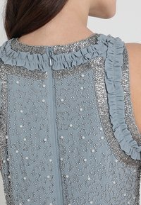 Ljusblå klänning med texturerad tyg som har paljetter och dekorationer, accentuerad med en volangkrage och en diskret dragkedja i ryggen.