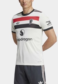 Hvit idrettsjersey med røde og svarte horisontale striper, strukturert stoff, svarte detaljer og logoer på forsiden og ermene.