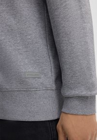 Szary sweatshirt o miękkiej fakturze, z prążkowanymi mankietami i małą szaro-skalowaną metką przy dolnym brzegu. Zaprojektowany z myślą o komforcie, o luźnym kroju.