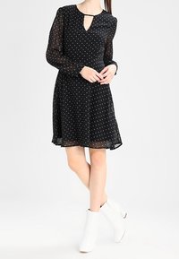 Robe noire à pois avec de longues manches transparentes, décolleté en trou de serrure et jupe évasée. Associée à des bottines blanches.