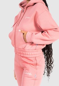 Sudadera rosa y joggers a juego en tela suave, con cremallera en el frente, bolsillo tipo canguro y detalles de logo bordado. Puños acanalados estilosos.