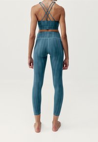 Žena stojící naboso, otočená zády k fotoaparátu, oblečená v tyrkysově modrých legínách na jógu a sportovní podprsence s křížovými popruhy na bílém pozadí.