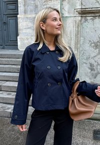 Tumši zila, dubultā jaciņa ar lielām pogām un brīvu piegriezumu. Modelis tur brūnu ādas roku somu, fonā redzami akmens kāpnes.