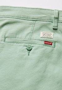Retro di un pantalone Levi's verde chiaro con chiusura a bottone, passante per cintura e due etichette del marchio: una bianca e una rossa.