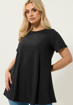 Blonde Frau trägt ein locker sitzendes, kurzärmeliges schwarzes Oberteil mit dezentem vertikalem Schimmer, posiert vor einem schlichten hellen Hintergrund.