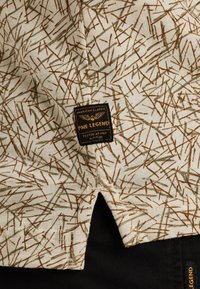 Camicetta a maniche corte in tessuto crema con un motivo di rametti marroni e verdi sparsi; etichetta nera con la scritta "PME Legend", a sottolineare la qualità.