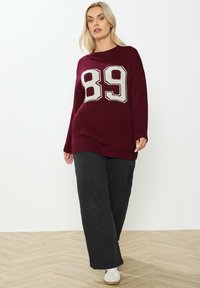Suéter burdeos con un gran diseño de punto en crema que dice "89", mangas largas y dobladillo acanalado. Combinado con pantalones negros de pierna ancha y zapatillas deportivas.