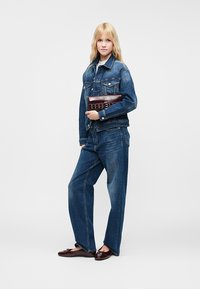Jeans jakna i široke traperice u srednje plavom denimu. Model drži smeđi clutch. Ravne, tamne cipele s malim mašnom.