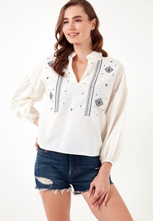Weiße Bluse mit navyblauer Stickerei, V-Ausschnitt und gepufften Ärmeln, kombiniert mit ausgewaschenen Denim-Shorts. Der Stoff hat eine strukturierte Oberfläche.