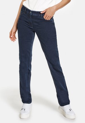 Femme portant un jean droit bleu foncé et des baskets blanches, debout avec une main dans la poche devant un fond uni.