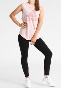 Helles rosa ärmelloses Shirt mit einem verblassten Adidas-Logo, kombiniert mit schwarzen Leggings und weißen Sportschuhen. Glatter Stoff, figurbetonter Schnitt.