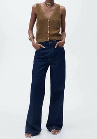 Brązowy dzianinowy kamizelka z dekoltem w kształcie litery V i guzikami, w połączeniu z szerokimi dżinsami w ciemnym kolorze. Podkreślona srebrną biżuterią i sandałami na odsłoniętych palcach.