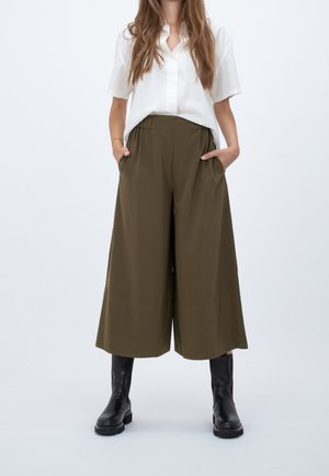 Woman wearing a white short-sleeve shirt, olive green wide-leg cropped pants, and black mid-calf boots, standing with hands in pockets.