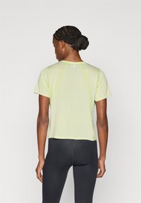 Αθλητική μπλούζα ανοιχτού πράσινου χρώματος με κοντά μανίκια, διαθέτει ανακλαστικές ραφές και άνετη εφαρμογή. Μαύροι κολάν φαίνονται στο κάτω μέρος.