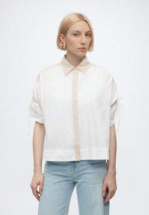 Mujer con cabello corto y rubio lleva una camisa blanca de botones extragrande con cuello beige y vaqueros azul claro, de pie contra un fondo blanco.