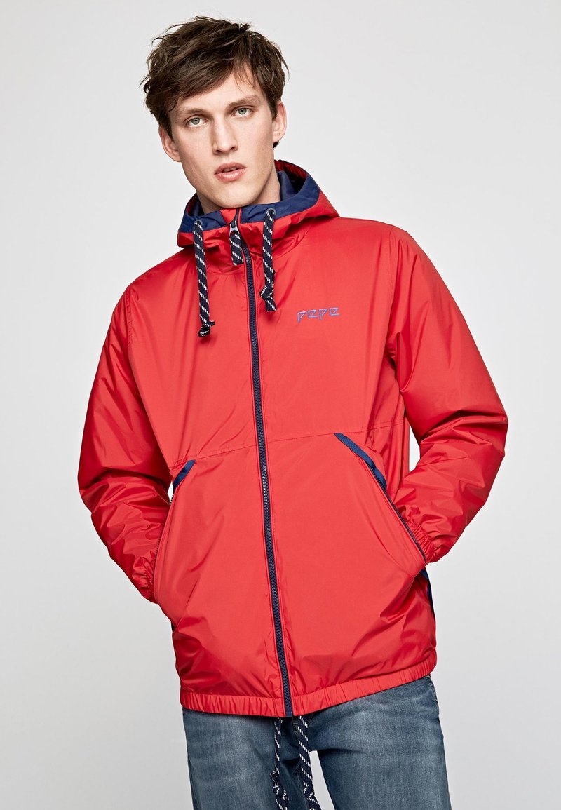 Veste imperméable rouge avec une capuche bleu marine, un cordon de serrage et une fermeture éclair à l'avant. Elle dispose de deux poches latérales et d'un logo sur la poitrine. Texture lisse.