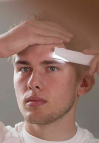 Man som håller en vit kam mot sin panna, med ljus som reflekteras från kammens kant, mot en neutral bakgrund.