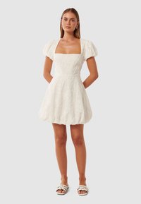 Weißes Kleid mit Puffärmeln und quadratischem Ausschnitt, mit strukturiertem Blumendesign, tailliertem Schnitt und ausgestellt Rock, kombiniert mit Sandalen mit offenen Zehen.