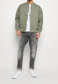 Olivgrön bomberjacka med ribbade muddar, tillsammans med urtvättade grå jeans och vita sneakers, och en grafisk vit t-shirt under.