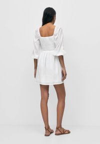 White, short puff-sleeve dress with a square back, textured fabric, fitted waist, and flared skirt. Worn with brown strappy sandals.