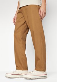Khaki trousers with a relaxed fit, woven texture, and side pockets; paired with beige sneakers featuring a textured sole and red accents.