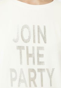 Witte katoenen T-shirt met textured zilveren letters die "JOIN THE PARTY" lezen, met een eenvoudig ronde halsontwerp en korte mouwen.