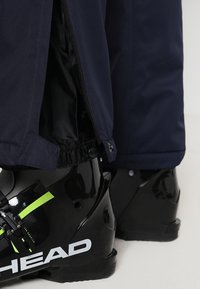 Bottes de ski noires avec le logo "HEAD", portées avec un pantalon de ski bleu foncé partiellement dézippé à la cheville, révélant la doublure intérieure et les détails de fixation.