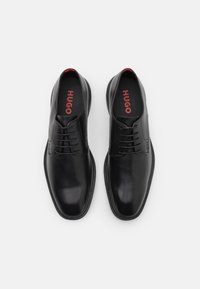 Chaussures habillées en cuir noir à finition lisse, avec un design à bout droit, six œillets et un logo rouge sur les semelles intérieures.