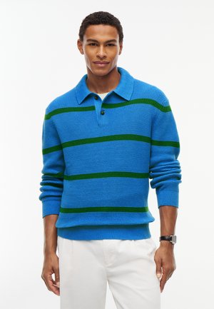 Mann trägt einen blauen Strickpullover mit grünen horizontalen Streifen und weiße Hose, steht vor einem schlichten weißen Hintergrund.
