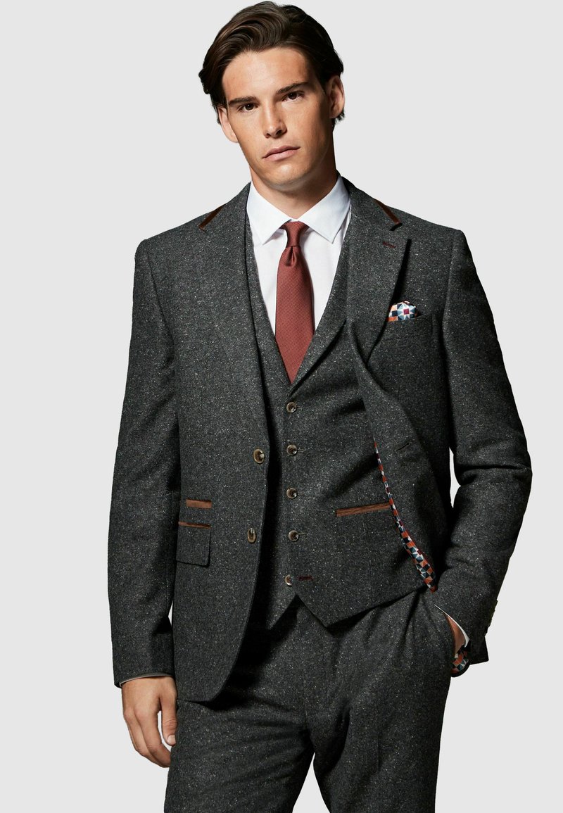 Hombre con traje de tres piezas gris oscuro con detalles marrones, camisa blanca y corbata color óxido, de pie con una mano en el bolsillo.